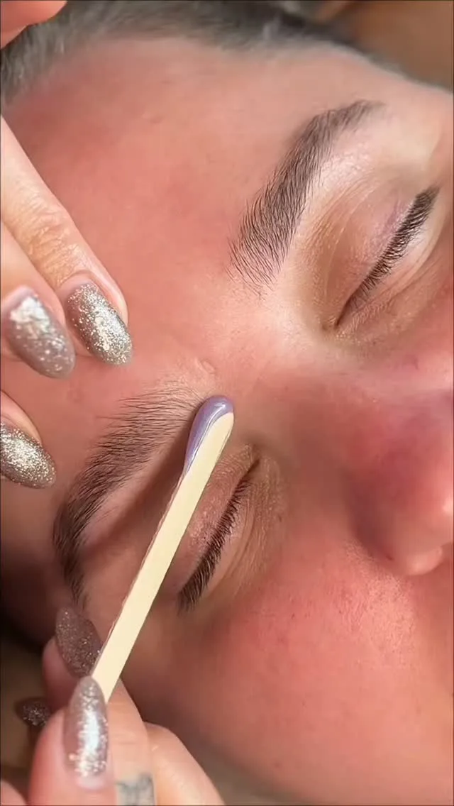 Wax stick applied along a brow, glitter-tipped nails — brow shaping process